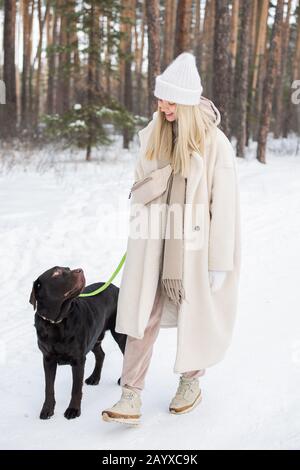 Horizontaler Schuss von jungen Frauen mit Kunstfellmantel, die mit ihrem schönen Hund am Wintertag im Freien verbringen Stockfoto