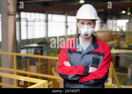 Horizontales Mittelporträt der ernsthaften Ingenieurin mit pritektivem Anzug, Helm und Maske, die mit gekreuzten Armen stehen Stockfoto