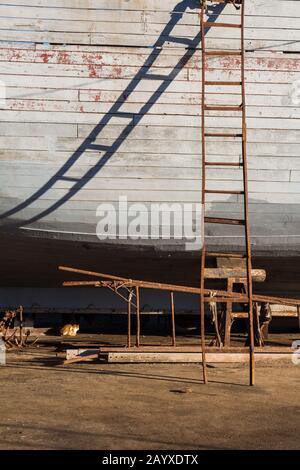 Seite eines Bootes aus dem Wasser im Hafen. Eiserne verrostete Leiter neben mit ihrem verdrehten Schatten auf dem Boot. Hafen von Essaouira, Marokko. Stockfoto