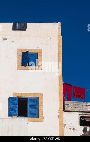 Ecke eines weißen Hauses mit blauen Blenden und gelben Rahmen. Tiefblauer Himmel. Rote Wäsche auf einem Seil auf einer Dachterrasse. Essaouira, Marokko. Stockfoto