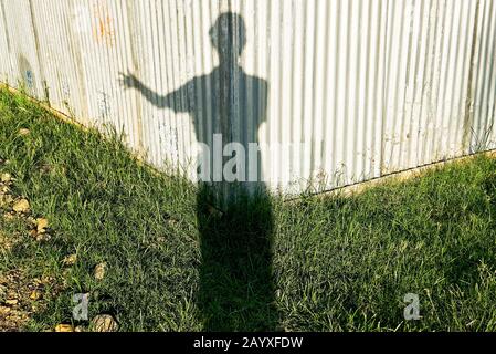 Ein Schatten eines jungen männlichen Menschen, der nur mit einer Hand grüßt und vor einer sonnenbeleuchteten Wellblechwand und einer Rasenfläche steht Stockfoto