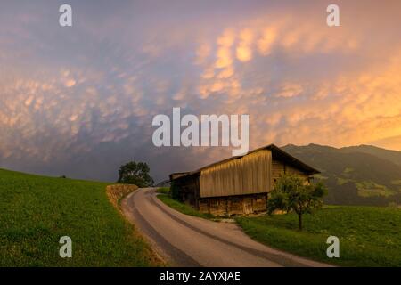 Atemberaubendes Landschaftsbild eines Cottages entlang einer Landstraße in den Bergen Österreichs. Bunte Sturmwolken als Hintergrund. Stockfoto