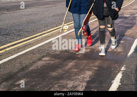 Zwei unerkennbare Teenager-Mädchen, die Skateboards tragen, die auf einem Radweg spazieren gehen. Eine trägt rote Ufer, die andere hat schwarze Jeans gerissen. Stockfoto