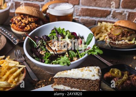 Auswahl an Gerichten, Salat mit Ziegenkäse hausgemachte Hamburger und pommes frites, Hühnerflügel mit jalapeños-Getränk und Kuchen auf dem Holztisch. Isola Stockfoto