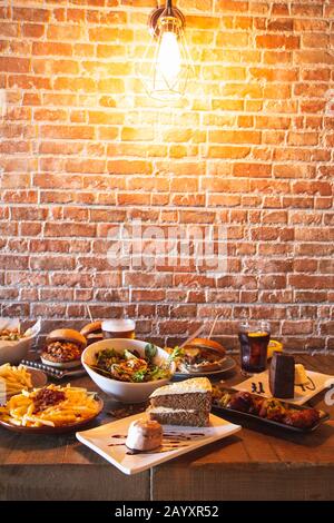 Verschiedene Menügerichte im Restaurant. Salat mit Ziegenkäse, hausgemachten Hamburgern und Pommes frites, Getränk, Hühnerflügel, Gemüsetempura und Kuchen auf dem Holz Stockfoto