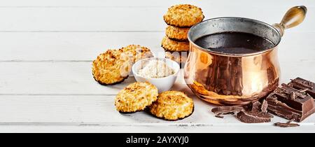 Kokosnuss-Makronen-Plätzchen mit Schokoladensockel neben einem Kupfertopf aus geschmolzener Schokolade und gehackter Süßigkeiten auf weißen Bretter in einem weiten Panorama Stockfoto