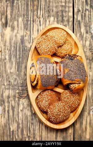 Hausgemachte frische Brötchen mit Mohnsamen und Plätzchen liegen auf einem ovalen Holzbrett auf einer alten Holzoberfläche in der Nähe eines vertikalen Bildes. Stockfoto