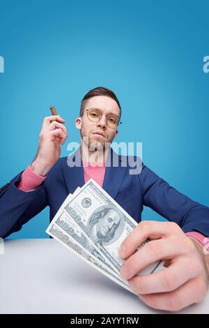 Porträt eines ernsthaften, selbstgerechten jungen bärtigen Mannes in einer Brille, die mit Geld am Tisch sitzt und Zigarre raucht Stockfoto