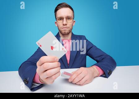 Porträt eines ernsthaften jungen Spielers in der Jacke am Tisch vor blauem Hintergrund sitzend und mit einer Ace Card Stockfoto