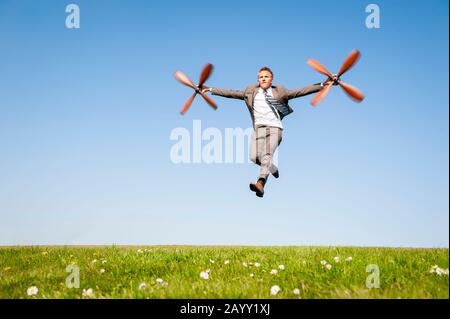 Geschäftsmann, der mit einem Satz drehender Flugzeugpropeller in der Luft über eine grüne Wiese fliegt Stockfoto