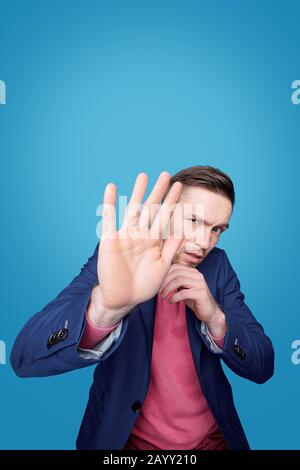 Porträt eines verängstigte jungen Mannes, der Stirn schaumelt und Gesicht hinter der Hand vor blauem Hintergrund versteckt Stockfoto