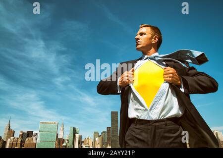 Starker junger Geschäftsmann, der seinen inneren Superhelden über der Skyline der Stadt im strahlend sonnigen blauen Himmel enthüllt Stockfoto