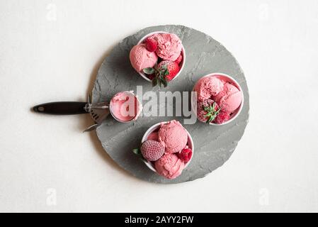 Hausgemachte Himbeeren Eiscreme in Schüsseln auf grauer runder Platte. Oben Blick auf Beeren Eis und gefrorene Früchte. Rote Eisschaufeln. Sommerdessert Stockfoto