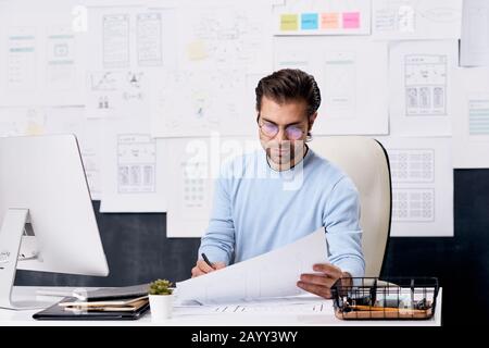 Horizontaler Schuss des jungen kaukasischen Mannes, der Brillen trägt, die am Büroschreibtisch sitzen und mit Papieren arbeiten Stockfoto