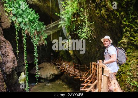 Junger starker lächelnder Mann in weißem Hut mit Rucksack am Höhleneingang. Wanderkonzept. Creek mit Wasserfall Stockfoto