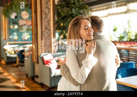 Fröhlicher junger Mann und Frau in Liebe begrüßen sich mit Umarmung im modernen Restaurant, horizontaler Mittelschuss Stockfoto
