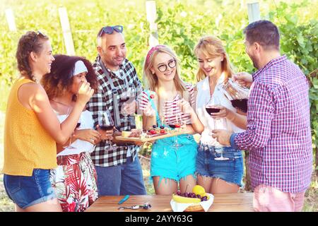 Junge, multiethnische Freunde, die im Freien Rotwein trinken, bei der Open-Air-Bar-B-que Party im ländlichen Restaurant mit Weinberg im Hinterhof Stockfoto