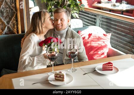 Horizontaler Schuss der schönen jungen Frau, die einen Haufen roter Rosen hält und ihren glücklichen Freund auf Wange küsst Stockfoto