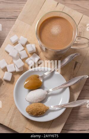 Auf einem Teelöffel liegen neben einem Glas Kaffee mit Milch Paukenzucker, Rohrzucker, loser Weißzucker und Buchweizenhonig. Stockfoto
