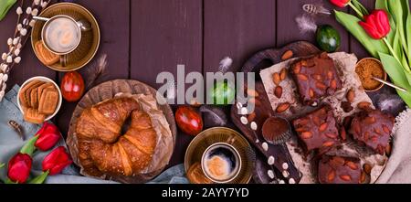Osterfrühstück mit aromatischem frischem Espresso, Croissants, Brownie-Kuchen, farbigen Eiern, roten Tulpen und Weide. Kaffee mit Gebäck, Blumen auf einem Holztisch. Ansicht von oben Stockfoto