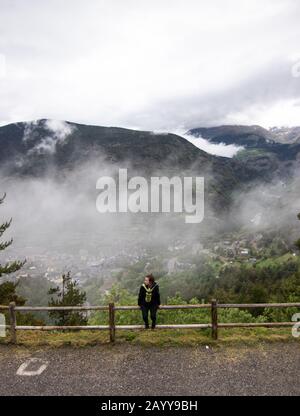 Touristenmädchen sitzt auf einem Holzzaun, während der Bergwanderung, mit Pyrenäenbergen im Hintergrund. Ein kleines Dorf von Encamp, in Andorra gelegen Stockfoto