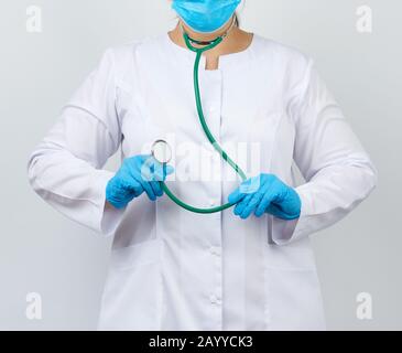 Medic Woman in einem weißen Mantel und Maske, mit blauen medizinischen Latexhandschuhen, hält ein grünes Stethoskop auf weißem Hintergrund, Studiofoto Stockfoto