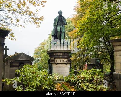 Alte Statue eines Mannes auf einem Kanonenteil eines Grabsteins auf einem monumentalen Friedhof mit Herbstbäumen und anderen Grabkapellen in einem grauen Himmel Stockfoto