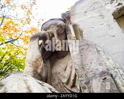 Nahaufnahme einer alten Statue einer trauernden Frau, die in weißen Stein gehauen ist, wobei ihr Kopf auf ihrer Hand einen Teil eines Grabmals unter einem Herbstbaum ruht Stockfoto