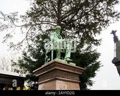 Alte Statue von alten Männern auf einem Stuhl an der Spitze eines alten Grab Denkmal unter dem Baum in einem monumentalen Friedhof sitzen Stockfoto