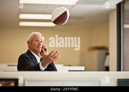 Leitender Geschäftsmann, der Fußball mit einer Hand in die Luft wirft, während er auf Handy spricht. Stockfoto