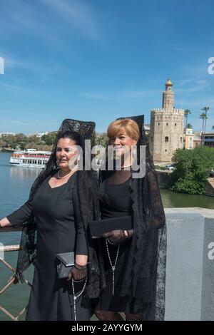 Zwei spanische Damen auf einer Brücke über den Fluss Guadalquivir mit dem Torre del Oro (Goldturm) im Hintergrund, die die traditionelle mantilla für Sem tragen Stockfoto