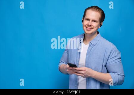 Ein Mann in einem blauen Hemd mit kabellosen Kopfhörern hält ein Smartphone und blickt auf die Kamera. Das Konzept der einfachen Kommunikation und der modernen Technik Stockfoto