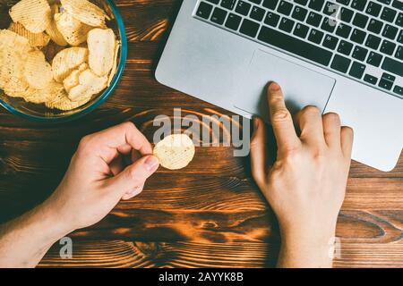 Glasplatte mit Spänen neben einem Laptop auf gebranntem Holzhintergrund. Der Mann hat Chips in den Händen und durchstöbert das Internet. Getont. Draufsicht. Nahaufnahme. Stockfoto