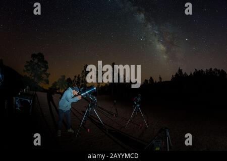 Junger Mann beobachtet Sternenhimmel durch ein Teleskop. In den Bergen, umgeben von Pinienwald im Hintergrund Nacht Landschaft mit bunten Milchstraße Stockfoto