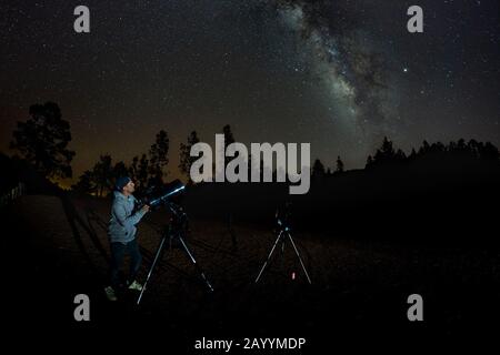 Junger Mann beobachtet Sternenhimmel durch ein Teleskop. In den Bergen, umgeben von Pinienwald im Hintergrund Nacht Landschaft mit bunten Milchstraße Stockfoto