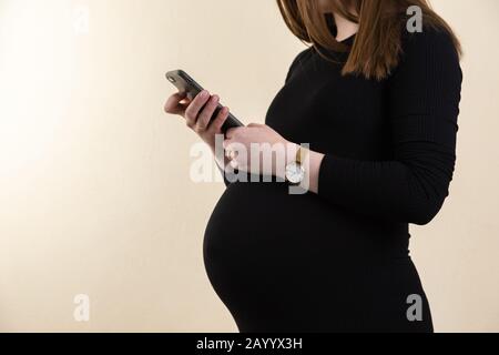 Die junge schwangere Frau in einem schwarzen Kleid hält ihren Bauch fest und sieht ihr Telefon an, während sie zu Hause bleibt. Stockfoto