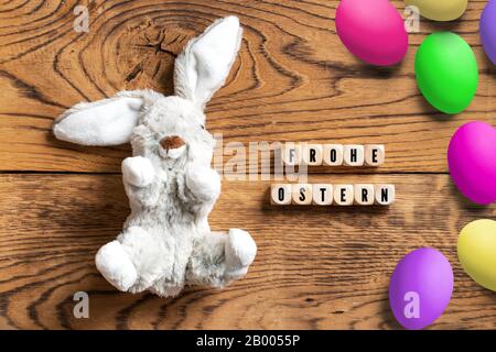 Gefülltes Häschen mit der Botschaft "Hapy Easter" auf Deutsch und vielen Eiern auf Holzhintergrund Stockfoto