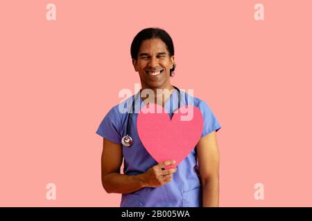 Studioporträt eines lächelnden männlichen Arztes oder einer Krankenschwester, die blaue Schrubben und Stethoskop trägt und die Herzaussparung hält Stockfoto
