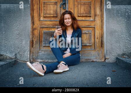 Junge Frau in Denimkleidung, die auf dem Boden über der altmodischen Holztür und dem Hintergrund der grauen Betonwand sitzt und Zuneigung in Phon sieht Stockfoto