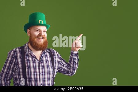 Alles gute zum St. Patrick's Day. Fetter komischer bärtiger rothaariger Mann in einem grünen Hut lächelnd zeigt einen Finger auf grünen Patrick's Tageshintergrund. Stockfoto