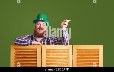 Alles gute zum St. Patrick's Day. Fetter komischer bärtiger rothaariger Mann in einem grünen Hut lächelnd zeigt einen Finger auf grünen Patrick's Tageshintergrund. Stockfoto