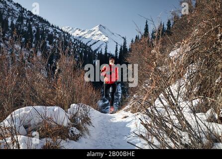 Der alte Mann mit grauem Bart und roter Jacke läuft im Winter in der Nähe der Berge. Skyrunning und wegweißendes Aktivitätskonzept. Stockfoto