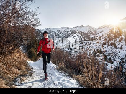 Der alte Mann mit grauem Bart und roter Jacke läuft im Winter bei Sonnenaufgang in der Nähe auf dem Weg in die Berge. Skyrunning und bahnbrechende Outdoor-Aktivitäten Stockfoto