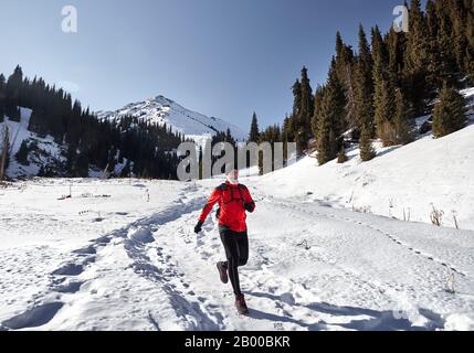 Der alte Mann mit grauem Bart und roter Jacke läuft in der Nähe der Berge im Winter an sonnigen Tagen. Skyrunning und wegweisende Outdoor-Acti Stockfoto