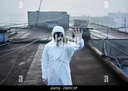 Forscher, der auf dem Dach eines Hochhauses steht und weißen Schutzanzug, Handschuhe und Gasmaske trägt, mit Blick auf die Kamera, gestiertes Stoppschild Stockfoto