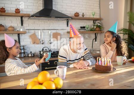 Bärtiger Mann in einem Geburtstagshut, der seinen Geburtstag mit seinen Enkeltöchtern feiert Stockfoto
