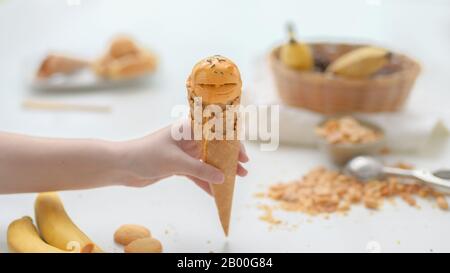Nahaufnahme eines Mädchens, das Erdnussbutter Bananen-Eiskegel mit verschwommener Erdnuss, Banane und Auffüllen auf weißem Tischhintergrund hält Stockfoto
