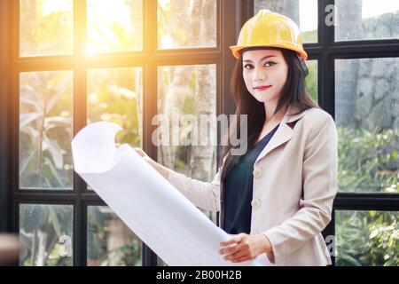 Porträt der schönen asiatischen Frau Architektenbauer mit gelbem Helm Harthut Studie Plan der Räume, ernsthafte Bauingenieurarbeiten w Stockfoto