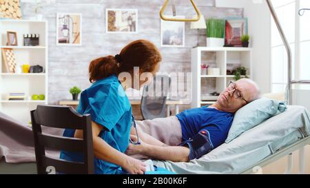 Weibliche Krankenschwester überprüft den Blutdruck des alten kranken Mannes. Der ältere Mann liegt in einem Krankenhausbett in einem hellen und gemütlichen Altenpflegeheim Stockfoto