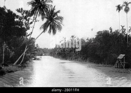 Thailand: Eine Kanalszene von außerhalb Bangkok, c.. 1900. Um die Wende des 20. Jahrhunderts waren die meisten Siamesen Reisbauern, die entlang der Wasserwege lebten und arbeiteten. Jeder Haushalt hatte ein Boot, von dem schätzungsweise 600,000 die Kanäle und Flüsse von Bangkok befuhren. Rudern wurde von der Rückseite des Bootes aus durchgeführt. Die meisten Häuser wurden aus Holz und Bambus gebaut und auf Stelzen mit einer Leiter zum Wasser gebaut. Auf dem rechten Ufer des Flusses stehen hohe, gerade Areca-Palmen, die Betelnüsse liefern; auf der linken Seite Kokosnusspalmen. Stockfoto